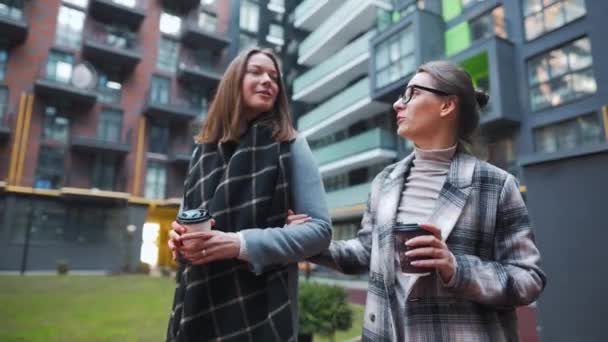 Dos mujeres felices caminando con café para llevar y hablando con interés entre ellas en el distrito de negocios. — Vídeo de stock