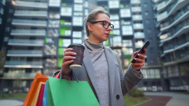 女性はビジネス街に立ち、テイクアウトコーヒー、ショッピングバッグを持ち、スマートフォンを使用しています。都市生活とデジタル技術の概念 — ストック動画