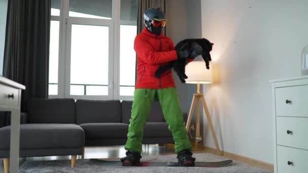 Весёлое видео. Человек, одетый сноубордистом, катается на сноуборде по ковру в уютной комнате. Он держит пушистую кошку на руках. В ожидании снежной зимы. Медленное движение — стоковое видео