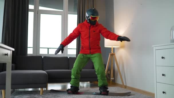 Hezké video. Muž oblečený jako snowboardista zobrazuje snowboarding na koberci v útulné místnosti. Čekám na zasněženou zimu. Zpomalený pohyb — Stock video