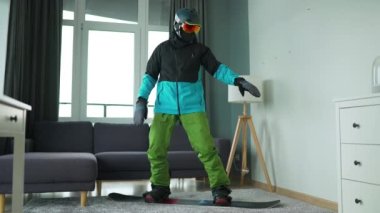 Eğlenceli video. Snowboard sporcusu gibi giyinmiş bir adam rahat bir odada halının üzerinde snowboard yaparken resmediyor. Karlı bir kış bekliyor