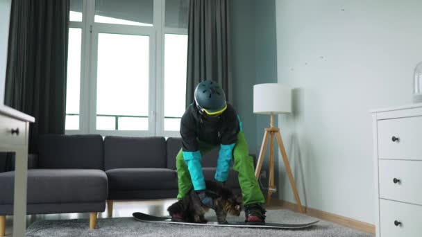 Весёлое видео. Человек, одетый сноубордистом, изображает сноубординг на ковре в уютной комнате с пушистым котом. В ожидании начала зимы — стоковое видео