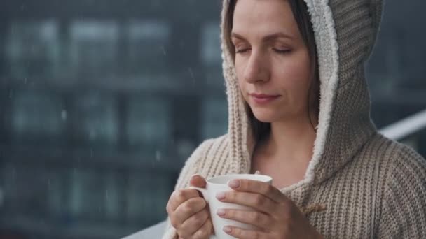 Kvinde opholder sig på altanen under snefald med kop varm kaffe eller te. Hun ser på snefnug og ånder i den kølige friske luft. – Stock-video