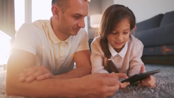 Baba ve kızı birlikte vakit geçiriyorlar ve evde oturma odasında yerde yatarken aile eğlencesi için tablet kullanıyorlar.. — Stok video