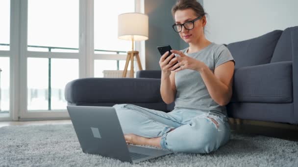 Alkalmanként öltözött nő ül szőnyegen laptoppal és okostelefonnal, és dolgozik a hangulatos szobában. Távoli munka az irodán kívül. — Stock videók