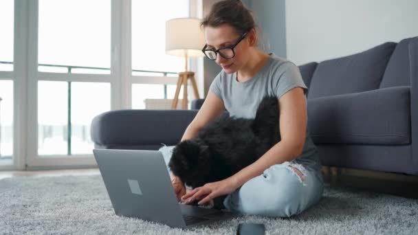 Mulher casualmente vestida com óculos senta-se em um tapete com um laptop e acaricia um gato preto fofo enquanto trabalha em uma sala aconchegante. — Vídeo de Stock