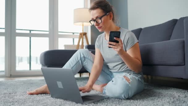 Wanita berpakaian santai duduk di karpet dengan laptop dan smartphone dan bekerja di kamar yang nyaman. Pekerjaan jarak jauh di luar kantor. — Stok Video