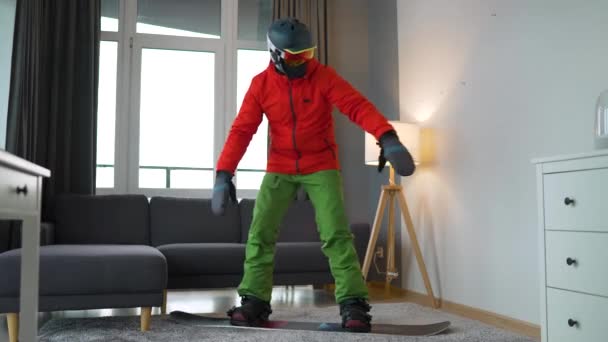 Hezké video. Muž oblečený jako snowboardista zobrazuje snowboarding na koberci v útulné místnosti. Čekání na sněhovou zimu — Stock video