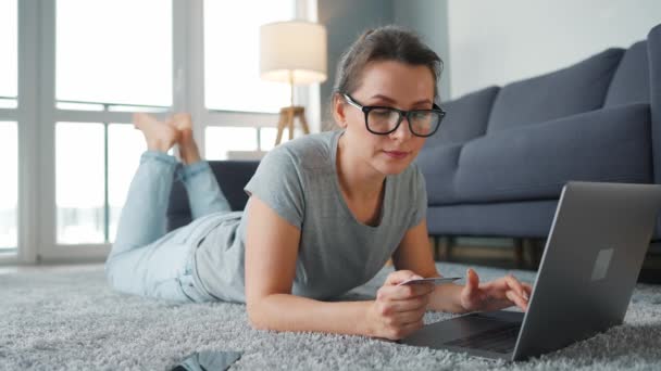 Mujer con gafas está tumbada en el suelo y hace una compra en línea con una tarjeta de crédito y un teléfono inteligente. Compras en línea, tecnología de estilo de vida — Vídeo de stock