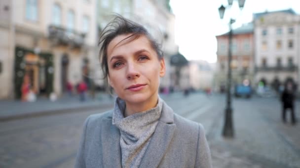 Portrait d'une femme caucasienne vêtue d'un manteau au milieu de la place de la vieille ville. Elle regarde sérieusement la caméra. Mouvement lent — Video