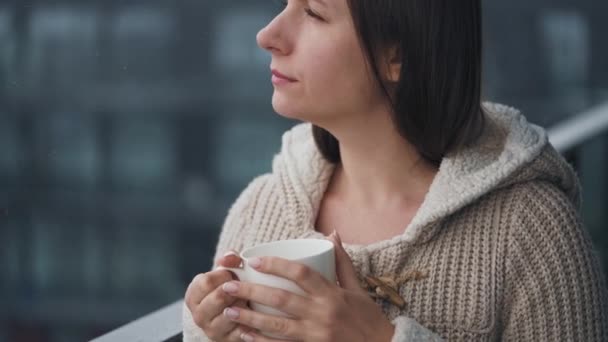 Vit kvinna stannar på balkongen under snöfall med kopp varmt kaffe eller te. Hon tittar på snöflingorna och andas i den frostiga friska luften. — Stockvideo