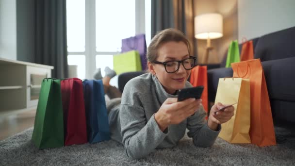 Lycklig kvinna ligger på golvet och gör ett online-köp med kreditkort och smartphone. Shoppingväskor. Onlineshopping, livsstilsteknik — Stockvideo