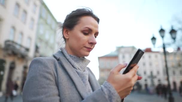 Žena v kabátě stojí uprostřed starého náměstí, používá smartphone a fotí. — Stock video