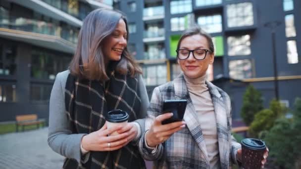 Dos mujeres felices caminando con café para llevar y hablando con interés entre ellas en el distrito de negocios. Movimiento lento — Vídeos de Stock