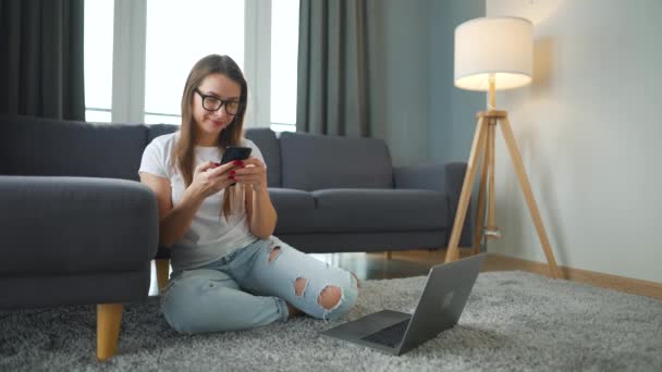 Mujer vestida casualmente sentada en la alfombra con portátil y teléfono inteligente y trabajando en una habitación acogedora. Trabajo remoto fuera de la oficina. — Vídeo de stock