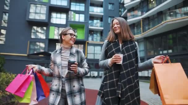 To lykkelige kvinner går med handleposer og kaffe etter en vellykket shopping og snakker med interesse seg imellom. Langsom bevegelse – stockvideo