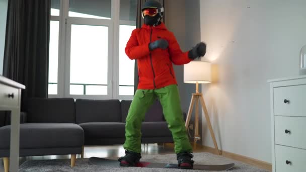 楽しいビデオだ。快適な部屋でカーペットの上のスノーボードに立っている間、男は喜んで踊り、楽しみを持ってスノーボーダーとして身を包んだ。雪の冬を待っている — ストック動画