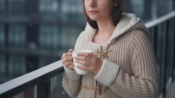 Donna rimane sul balcone durante la nevicata con una tazza di caffè caldo o tè. Guarda i fiocchi di neve e respira l'aria fresca gelida. — Video Stock