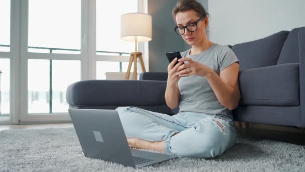 Alkalmanként öltözött nő ül szőnyegen laptoppal és okostelefonnal, és dolgozik a hangulatos szobában. Távoli munka az irodán kívül. — Stock videók