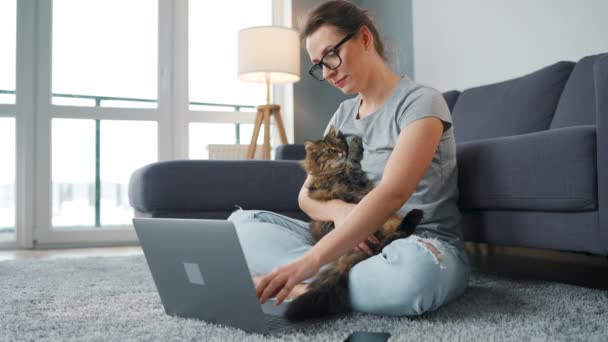 Mulher casualmente vestida senta-se em um tapete com um laptop, segura os joelhos e acaricia um gato fofo e trabalha em uma sala aconchegante. Trabalho remoto fora do escritório. — Vídeo de Stock