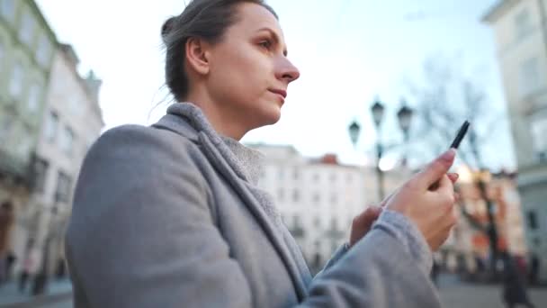 Žena v kabátě stojí uprostřed starého náměstí, používá smartphone a fotí. — Stock video