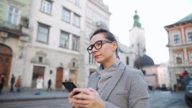 Gözlüklü bir kadın eski bir caddede yürürken ve akıllı telefon kullanırken.