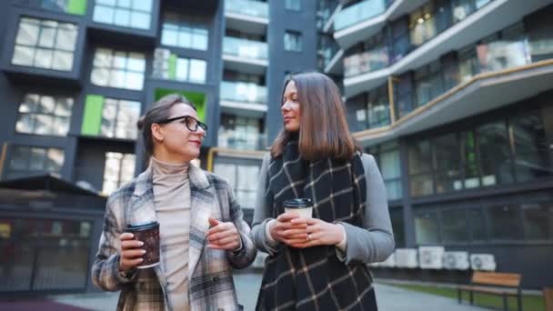 Dwie szczęśliwe kobiety spacerujące z kawą na wynos i rozmawiające z zainteresowaniem w dzielnicy biznesowej. Zwolniony ruch — Wideo stockowe