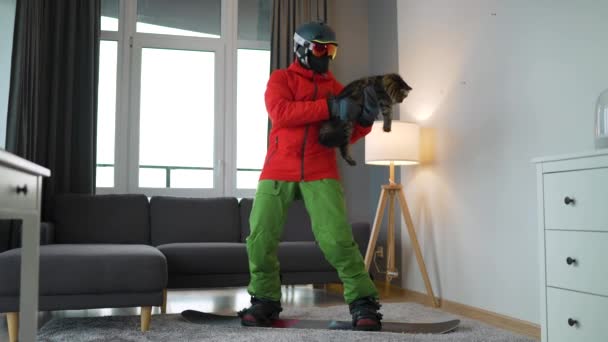 Rolig video. Man klädd som en snowboardåkare åker snowboard på en matta i ett mysigt rum. Han håller en fluffig katt i sina armar. Väntar på en snöig vinter. Långsamma rörelser — Stockvideo