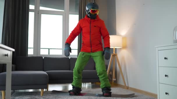 Vídeo divertido. Hombre vestido de snowboarder representa el snowboard en una alfombra en una habitación acogedora. Esperando un invierno nevado. Movimiento lento — Vídeos de Stock
