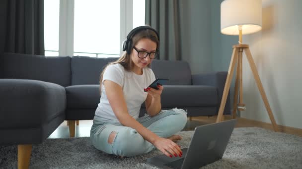 Casualmente mujer vestida con auriculares está sentado en la alfombra con el ordenador portátil y el teléfono inteligente y trabajando en una habitación acogedora. Trabajo remoto fuera de la oficina. — Vídeos de Stock