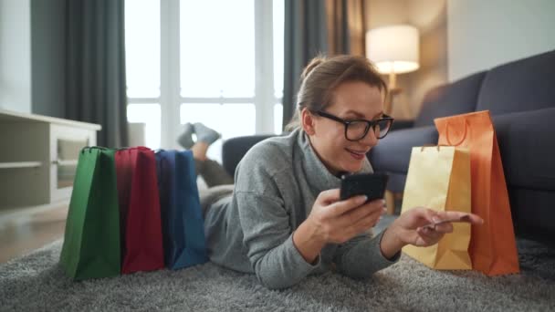 Mujer feliz está tirada en el suelo y hace una compra en línea con una tarjeta de crédito y un teléfono inteligente. Bolsas de compras alrededor. Compras en línea, tecnología de estilo de vida — Vídeos de Stock