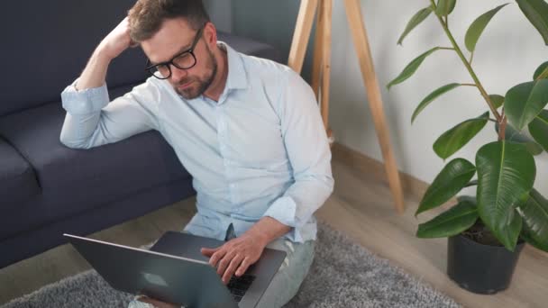 Man med glasögon sitter på matta med laptop och smartphone och arbetar i mysiga rum. Fjärrarbete utanför kontoret. — Stockvideo