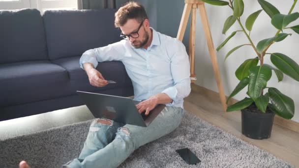 Hombre con gafas sentado en el suelo y hace una compra en línea con una tarjeta de crédito y un ordenador portátil. Compras en línea, tecnología de estilo de vida — Vídeo de stock