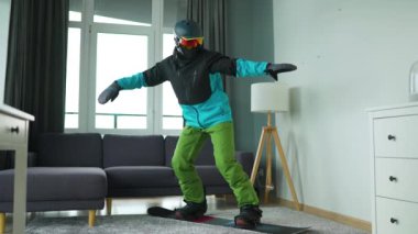 Eğlenceli video. Snowboard sporcusu gibi giyinmiş bir adam rahat bir odada halının üzerinde snowboard yaparken resmediyor. Karlı bir kış bekliyor. Yavaş çekim