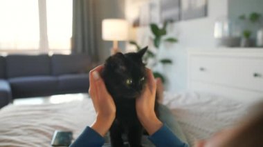 Kadın, rahat bir odada, yatakta uzanmış siyah bir kediyi okşuyor..