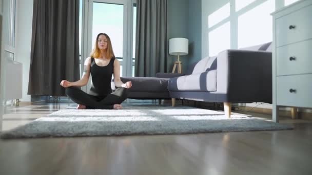 Mujer joven caucásica en mono negro sentada en posición de loto y meditando, yoga en casa. — Vídeos de Stock