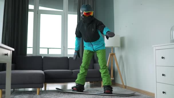 Vídeo divertido. Hombre vestido de snowboarder representa el snowboard en una alfombra en una habitación acogedora. Esperando un invierno nevado. Movimiento lento — Vídeos de Stock