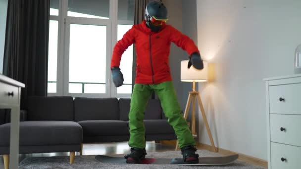 Vídeo divertido. Hombre vestido de snowboarder representa el snowboard en una alfombra en una habitación acogedora. Esperando un invierno nevado. Movimiento lento — Vídeo de stock