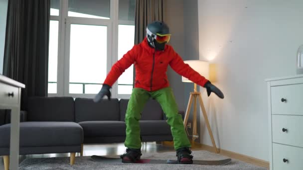 Hezké video. Muž oblečený jako snowboardista zobrazuje snowboarding na koberci v útulné místnosti. Čekám na zasněženou zimu. Zpomalený pohyb — Stock video