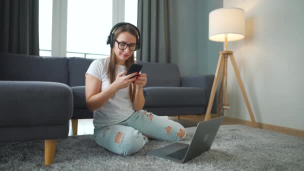 Alkalmanként öltözött nő fülhallgatóval ül a szőnyegen laptop és okostelefon és dolgozik a hangulatos szobában. Távoli munka az irodán kívül. — Stock videók