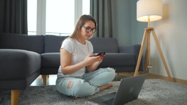 Nieformalnie ubrana kobieta siedzi na dywanie z laptopem i smartfonem i pracuje w przytulnym pokoju. Praca zdalna poza biurem. — Wideo stockowe