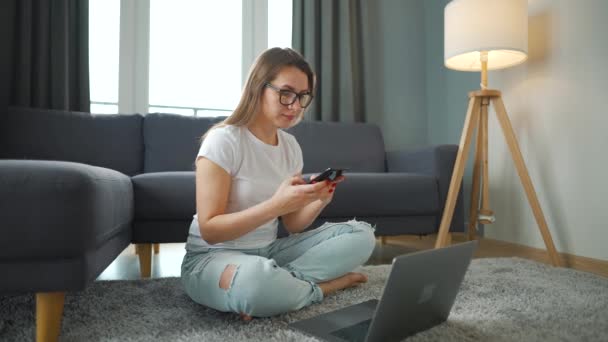 Mulher casualmente vestida está sentada no tapete com laptop e smartphone e trabalhando em quarto acolhedor. Trabalho remoto fora do escritório. — Vídeo de Stock