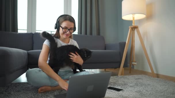 Wanita berpakaian santai duduk di karpet dengan laptop dan bekerja dari jarak jauh di kamar yang nyaman. Kucing berbulu hitam datang ke lututnya dan dia membelainya — Stok Video