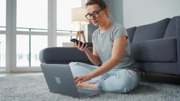 Lässig gekleidete Frau sitzt auf Teppich mit Laptop und Smartphone und arbeitet in gemütlichem Raum. Fernarbeit außerhalb des Büros. — Stockvideo