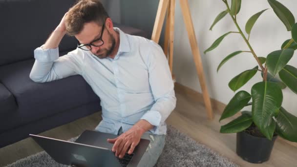 Hombre con gafas sentado en la alfombra con ordenador portátil y trabajando en una habitación acogedora. Trabajo remoto fuera de la oficina. — Vídeos de Stock