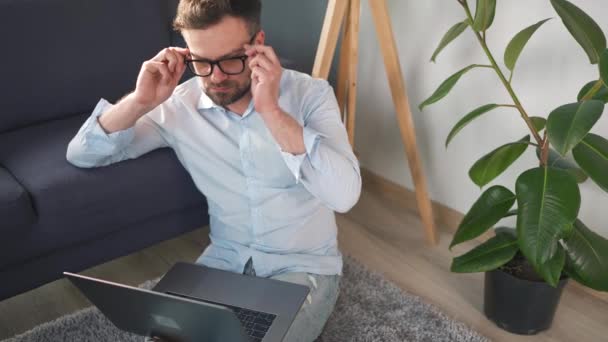 Hombre con gafas sentado en la alfombra con ordenador portátil y trabajando en una habitación acogedora. Trabajo remoto fuera de la oficina. — Vídeos de Stock