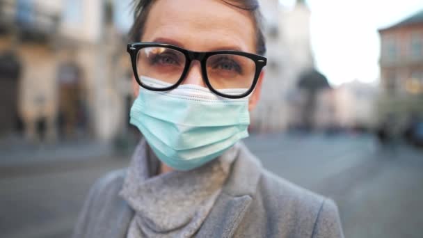 Covid-19 koronavirüsünün pandemik koruması. Ceketli, gözlüklü ve koruyucu sağlık maskeli bir kadın portresi. Meydanın ortasında duruyor. Yavaş çekim — Stok video