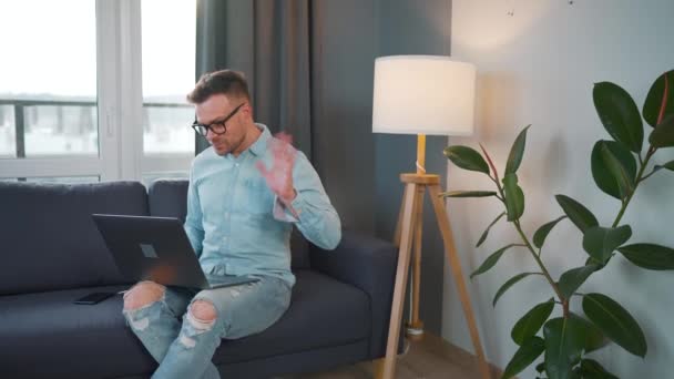 Hombre vestido casual con gafas sentado en el sofá y el uso de la computadora portátil para hacer una videollamada desde casa en un apartamento moderno — Vídeos de Stock