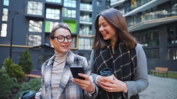 Δύο ευτυχισμένες γυναίκες περπατούν με καφέ και μιλάνε με ενδιαφέρον μεταξύ τους στην επιχειρηματική περιοχή. Αργή κίνηση — Αρχείο Βίντεο