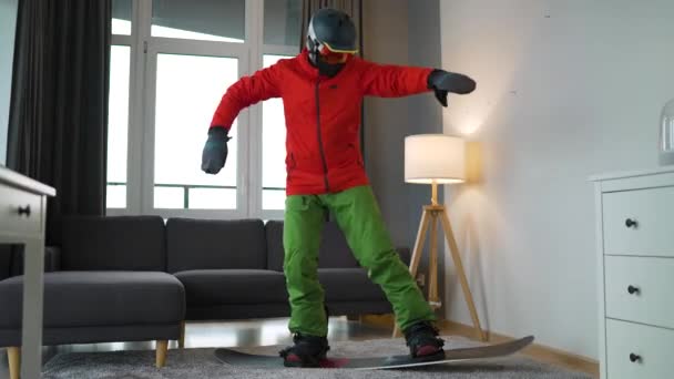 Vídeo divertido. Hombre vestido de snowboarder representa el snowboard en una alfombra en una habitación acogedora. Esperando un invierno nevado. — Vídeos de Stock